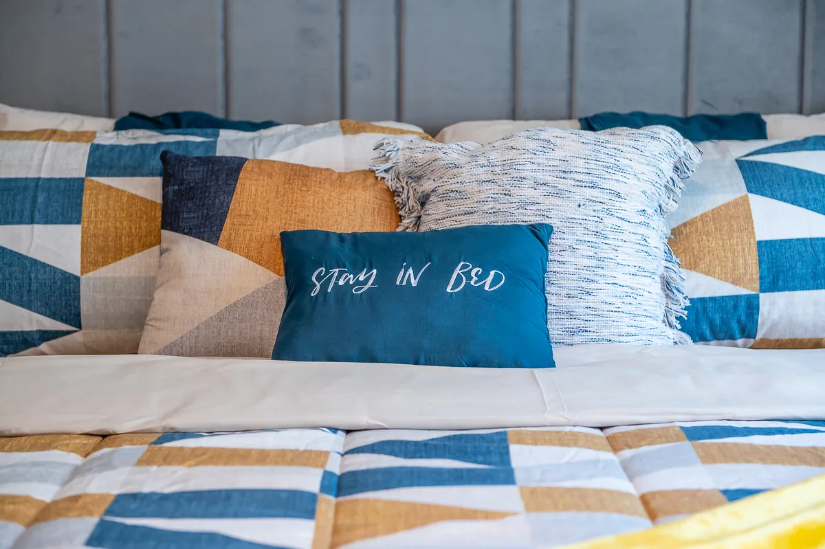 A close-up of a bed adorned with colorful throw pillows and geometric patterned bedding, along with a calming blue accent pillow featuring text. The scene feels inviting and fresh, ideal for relaxing or reading.