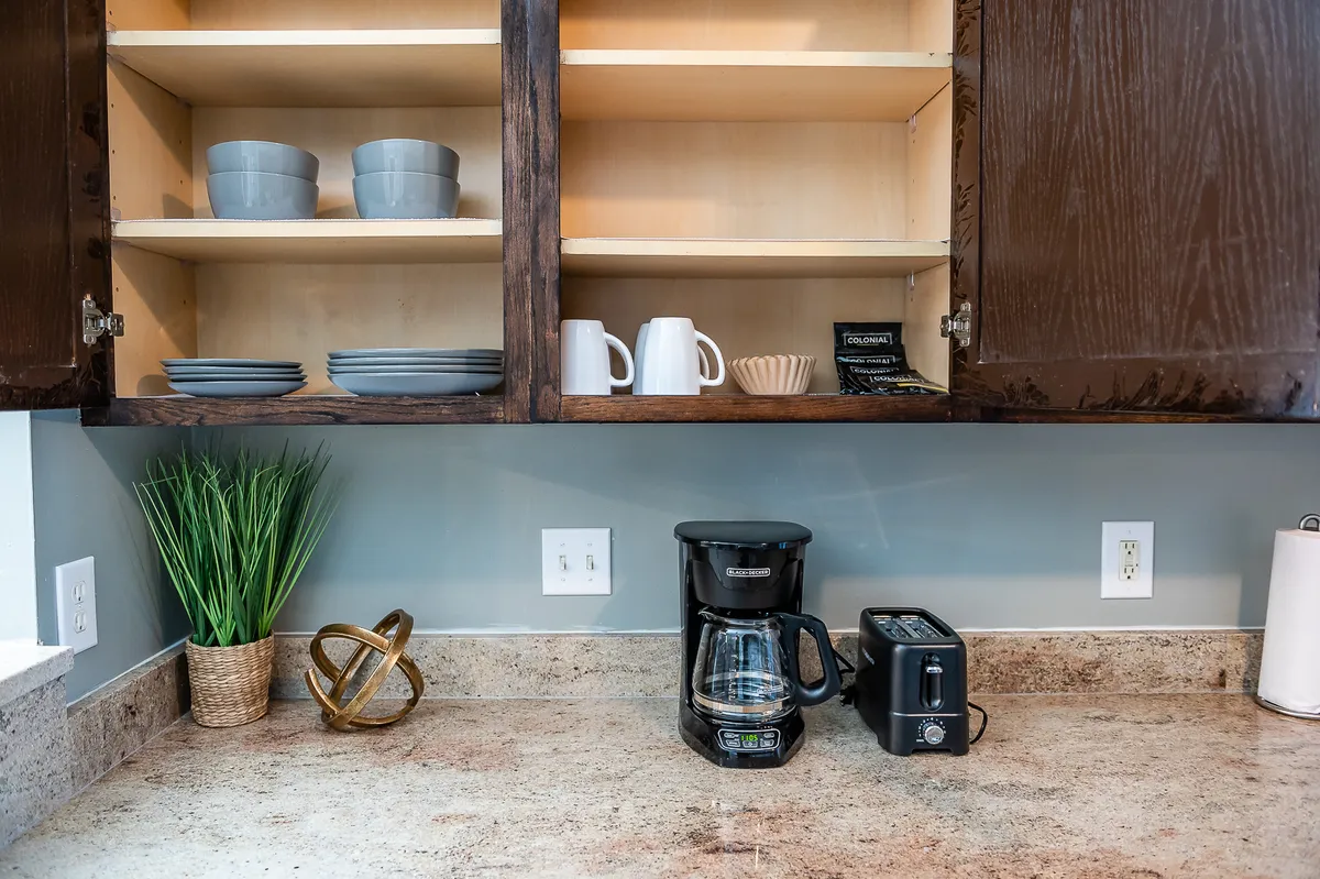 A cozy kitchen scene featuring a coffee maker, toaster, stacks of plates, a decorative bowl, and a green plant. The breakfast setting and natural elements create an inviting atmosphere for morning routines and shared stories.