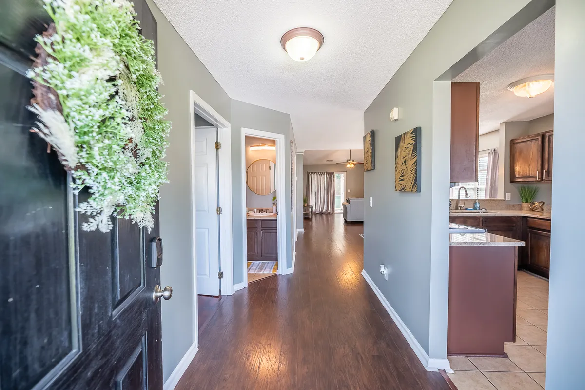 This image shows a warm and inviting entryway featuring a front door adorned with a wreath, a hallway that seamlessly leads into the kitchen and living space, and tasteful wall art. The perspective highlights the home's open layout and immediate access to living areas as soon as you enter.