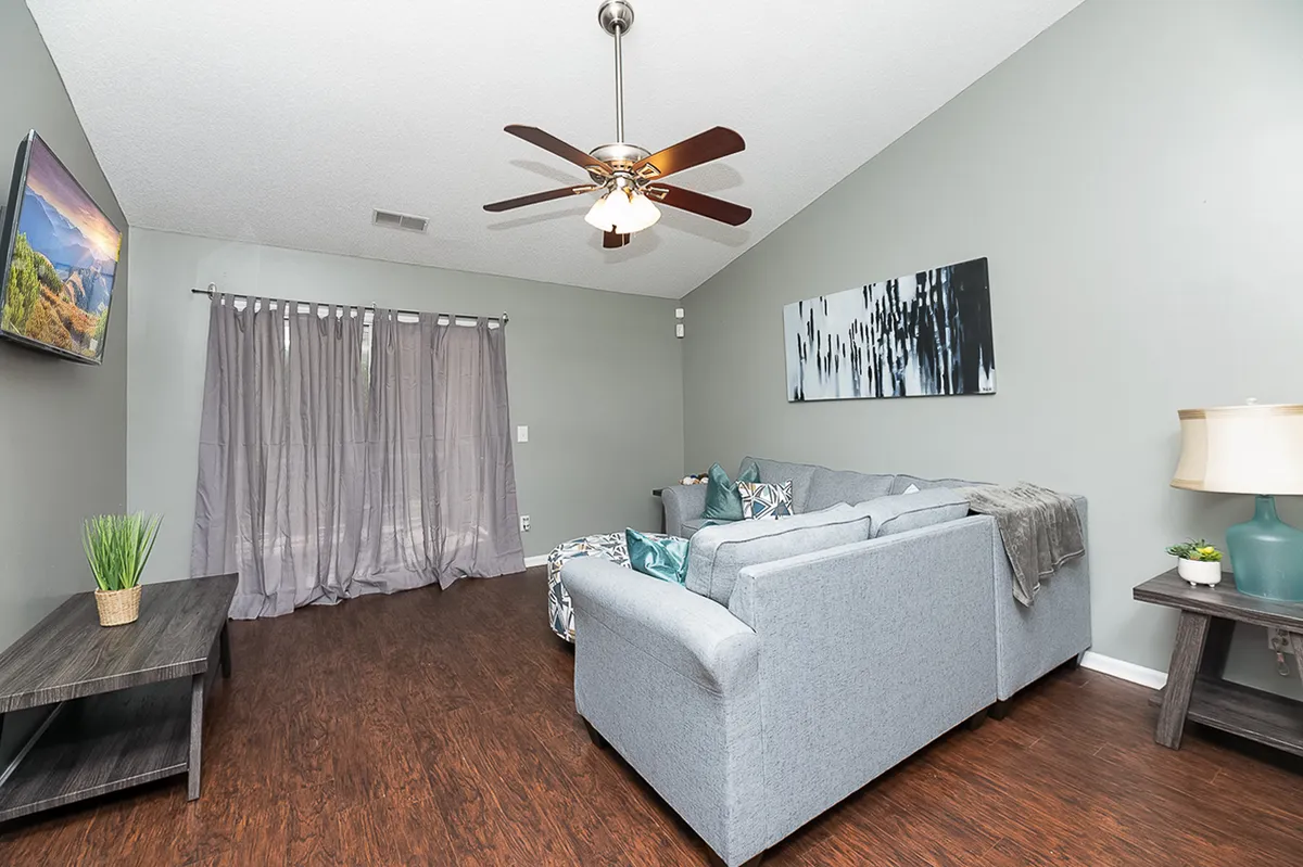 A cozy living room featuring a plush sofa, ceiling fan, stylish wall artwork, flowing curtains, and a coffee table. The layout is open and inviting, ideal for relaxing, socializing, or watching TV.