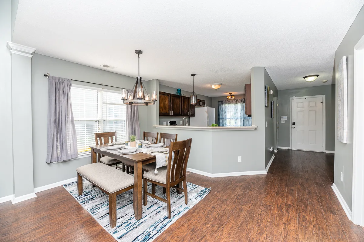 A bright, open dining area with a kitchen in the background, filled with natural light from a large window. The scene is set for family meals, entertaining, or casual dining, showcasing both practicality and warmth in communal living spaces.