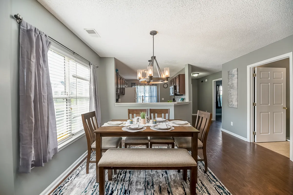The image showcases a bright, inviting dining area featuring a stylish dining table beneath an elegant chandelier, with plenty of natural light pouring in from a large window. The open concept seamlessly connects the dining space to a modern kitchen, creating the perfect setting for family meals, entertaining friends, or enjoying a relaxed weekend brunch.