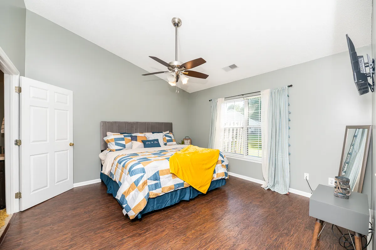 A comfortable bedroom featuring a large window with curtains, a cozy bed dressed in colorful bedding, a ceiling fan, and a wall-mounted television. The natural light creates a welcoming environment suitable for relaxing or catching up on work remotely.