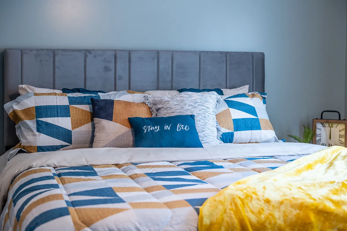 A bedroom scene featuring a bed with geometric-patterned bedding, decorative pillows, a soft headboard, and a yellow throw blanket. The mood is warm and inviting, emphasizing comfort and coziness.