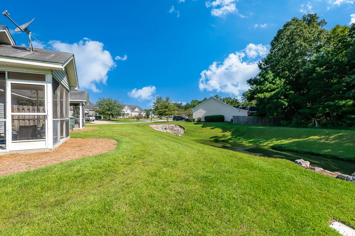 A spacious green lawn is bordered by a tranquil water feature, with mature trees and neighboring homes in the background. The area looks ideal for relaxing outdoors, having family picnics, or letting children play. The natural elements and open space evoke a sense of peace and community.