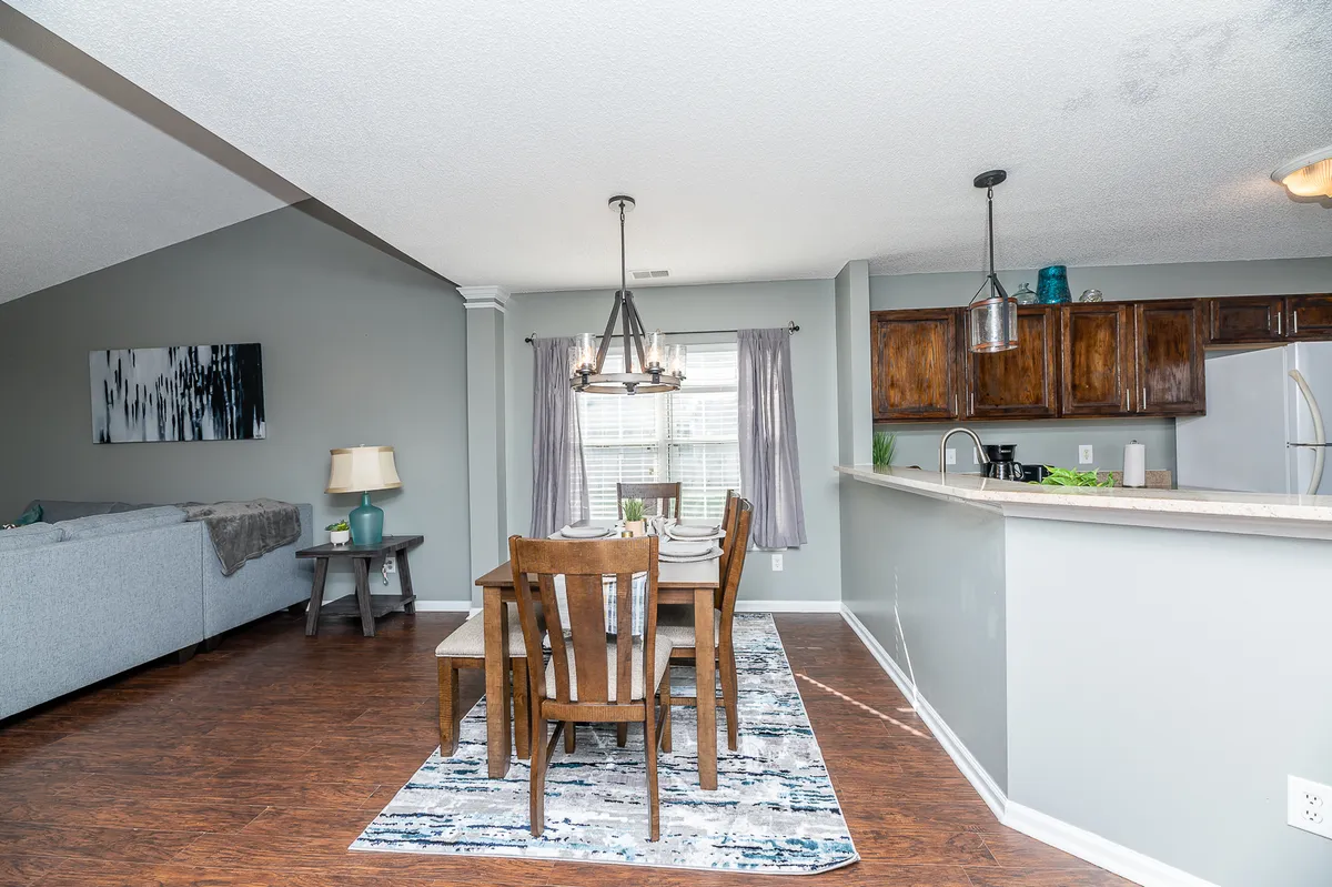 A spacious, open-concept area that seamlessly connects the dining table, kitchen, and visible sofa in the living area, all bathed in warm chandelier lighting. The setup evokes a sense of togetherness and easy conversation between spaces.